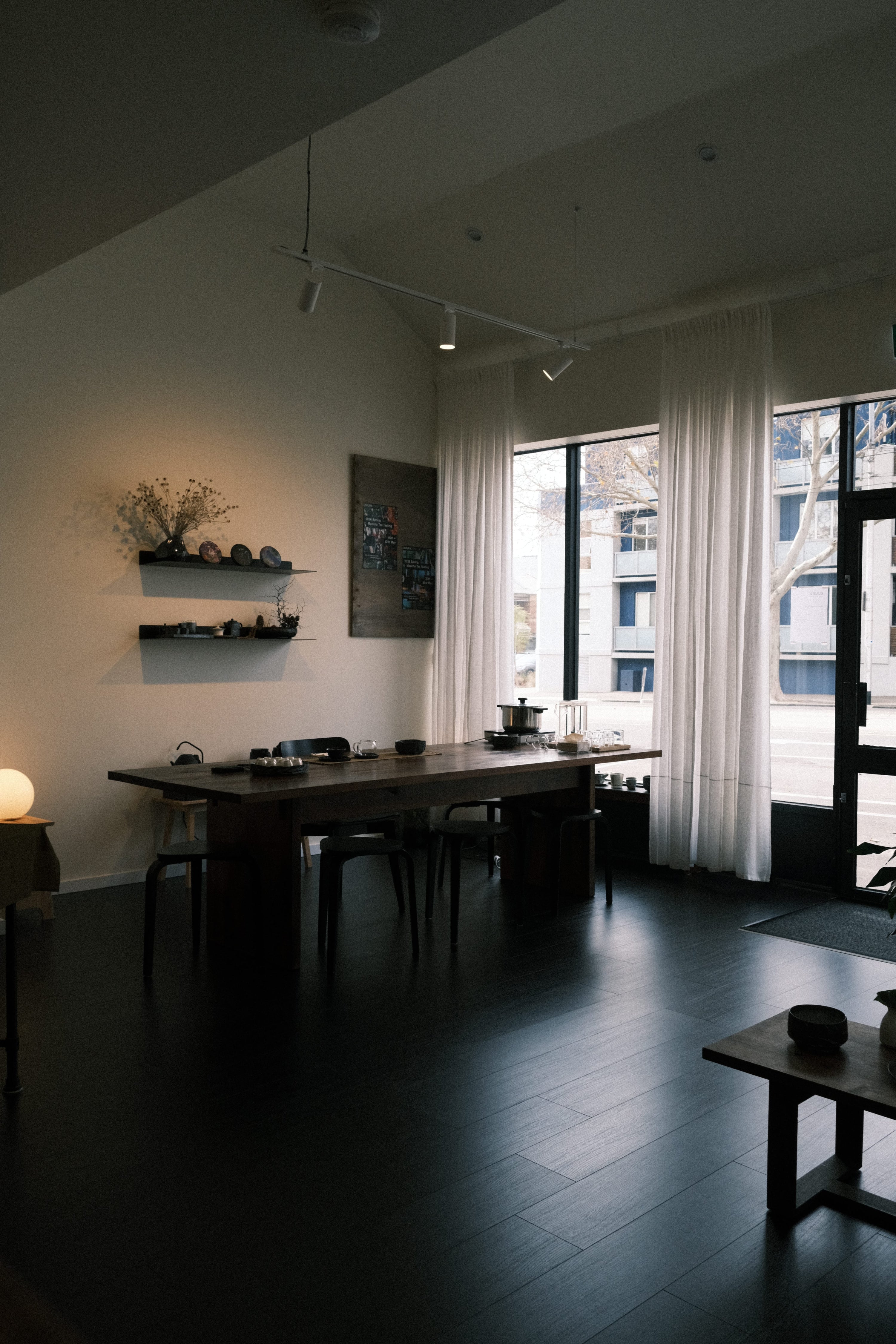 A dark wooden tea table with matching chairs in a modern, dimly lit room with white walls, sheer curtains, and dark hardwood floors.
